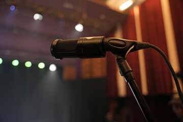 Obraz premium A black microphone on a stand, positioned on a stage with warm lighting and blurred background of curtains and spotlights.