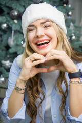 Joyful Woman in Fluffy White Winter Hat Laughing and Making a Heart Shape with Her Hands near a Christmas Tree
