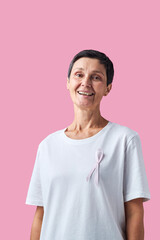 Portrait of middle aged Caucasian woman smiling and standing against pink background wearing shirt with pink ribbon symbolizing breast cancer awareness