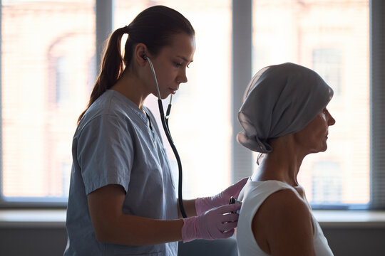 Caucasian female healthcare worker examining back of middle aged Caucasian woman wearing headscarf with stethoscope in medical setting, cancer patient receiving checkup - Powered by Adobe