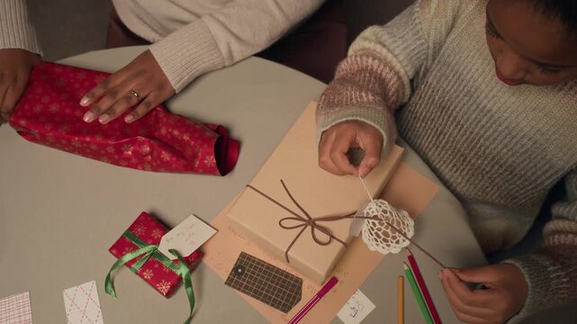 High angle shot of focused Black girl child decorating gift box with twine and baubles while preparing presents for Christmas party sitting at table with family in living room