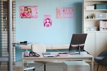 Empty medical office showing modern computer monitor, stethoscope, documents on desk, breast cancer awareness posters on blue wall, exam table in background, healthcare setting