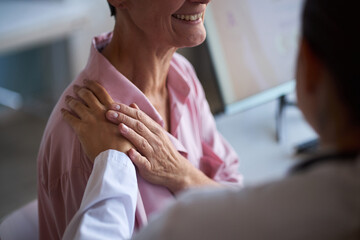 Middle aged Caucasian woman sitting in medical office receiving comforting touch from female doctor during cancer consultation, both women partially visible, focus on supportive gesture