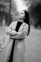 Fototapeta premium Asian woman in a stylish coat stands confidently on a tree-lined path, showcasing a serene expression and elegant posture, embodying grace and poise in a tranquil outdoor setting