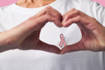 Middle aged Caucasian woman forming heart shape with hands in front of chest, wearing shirt with pink ribbon symbol, representing breast cancer awareness and support