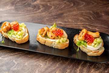 Gourmet bruschetta with grilled shrimp, red caviar, cucumber, and cream cheese on toasted bread, garnished with microgreens. Elegant appetizer on black plate, top view.
