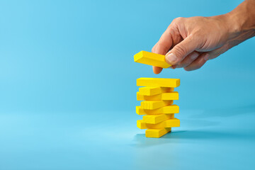 Building a yellow stacking game with hand balancing blocks on a blue background to create a tall tower for fun and creativity, copy space.