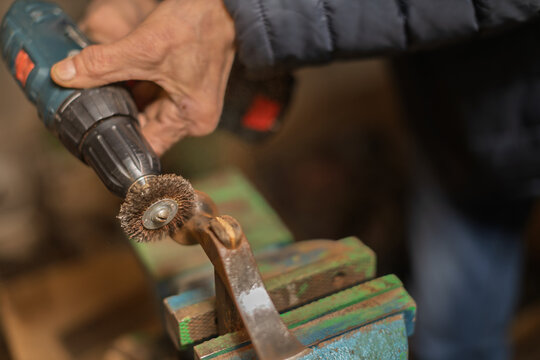 A person uses a power drill with a wire brush attachment to clean a metal object secured in a vise.
