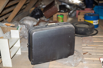 A dusty black suitcase sits on a wooden floor in a cluttered attic filled with various stored items...