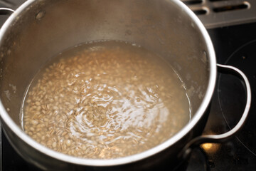 Pearl barley is shown boiling in a metal pot on a stovetop, with visible bubbles and ripples indicating active cooking.