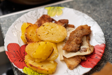 A plate of golden-brown roasted potato slices seasoned with caraway seeds and fried meat topped with melted cheese is presented on a decorative plate.