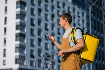 Male bike courier delivering food in urban city, wearing a yellow thermal backpack and checking a smartphone for navigation or order details, working in the gig economy