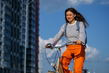 Young woman cycling a city bike along an urban street, carrying a blue backpack, wearing a striped...