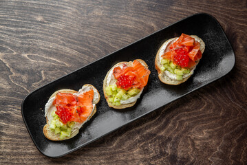 Gourmet bruschetta with cream cheese, cucumber, smoked salmon, and red caviar on toasted bread, served on a black plate. Elegant appetizer for upscale dining or catering.