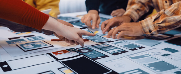A diverse group of individuals engages in collaborative brainstorming over colorful paper mockups, focusing on interface design strategies in a modern office environment. SACTR