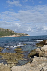 Rocky shoreline and turquoise Mediterranean waters of Cala Agulla Mallorca with lush green hills under a partly cloudy sky
