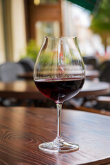 A beautifully shaped glass filled with deep red wine sits on a wooden table outside a bustling cafe