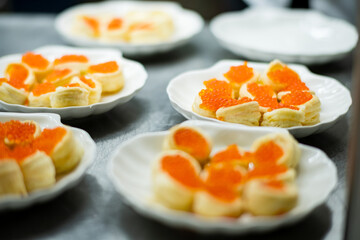 Savory pastries sit on white plates, adorned with bright orange caviar, in a lively kitchen space