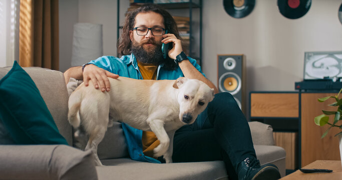 A relaxed man speaks on the phone with a family member while gently petting his dog, who is waiting near the sofa.