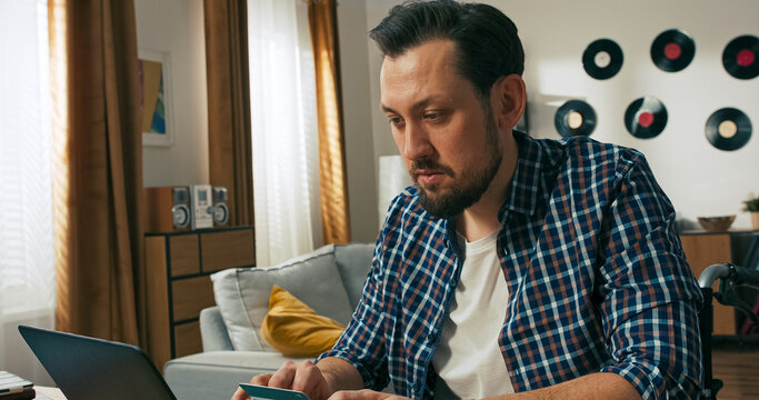 A focused man who uses a wheelchair holds a credit card and types its number into his laptop. He is young, successful, and visibly pleased with the ease of managing payments online. - Powered by Adobe