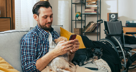 A middle-aged man with a disability sits on a couch using his phone, surrounded by his dogs, enjoying a peaceful moment at home with loyal companions by his side.