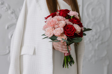 Close-up of a woman in a white dress and blazer holding a beautiful mixed bouquet of deep red roses, blush roses, and pink carnations. Elegant and romantic style