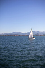 Fototapeta premium A lone sailboat sails on the blue water of the Bay of Santander on a sunny day, with the coastline and mountains visible in the distance.