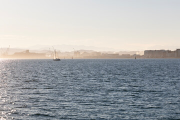 A lone sailboat sails on the blue water of the Bay of Santander on a sunny day, with the coastline and mountains visible in the distance.