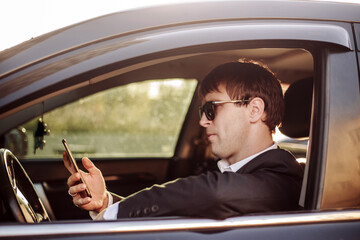 Businessman in a suit and glasses sits in car, looking at and talking on the phone.