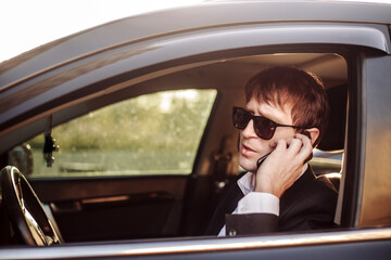Businessman in suit and glasses sits in car, looking at and talking on the phone.