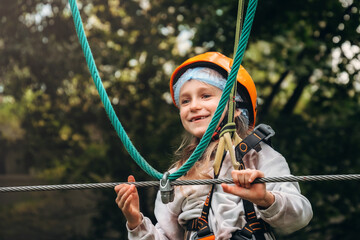 Smiling young girl wearing a helmet navigates a rope course in an adventure park. Her expression shows excitement and enjoyment during the activity, surrounded by lush greenery.