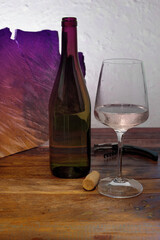 A rustic and minimalist still life of a red wine bottle and glass on a wooden table against a white textured wall.