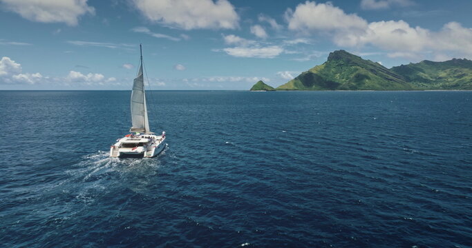 Fiji, Kuata island: White catamaran with tourists sailing blue ocean water near tropical island, under bright sunny sky. Remote wild nature paradise, exotic summer travel. Aerial drone flight