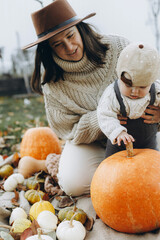 Stylish happy mother and adorable baby son playing on cozy blanket with pumpkin and autumn leaves....