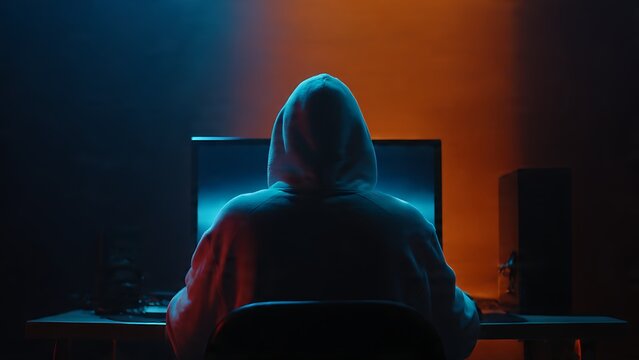 A person sits in front of a computer monitor, likely engaged in covert or suspicious activity, shrouded in shadow