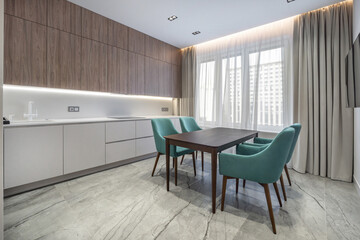 A bright and sleek kitchen features wooden cabinets and a dining table surrounded by green chairs. Large windows illuminate the space, highlighting modern design elements