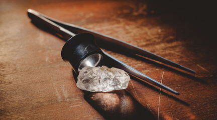 Raw uncut diamond with jeweler loupe and tweezers on wooden surface