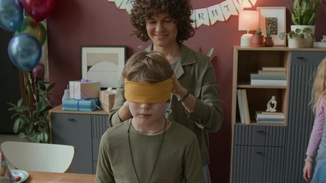 Woman putting blindfold over eyes of boy in cowboy costume before playing blindmans buff with friends during birthday party