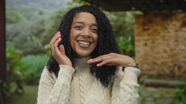 African american woman wearing cable knit sweater smiles and frames face with hands in lush green park; joy confidence.