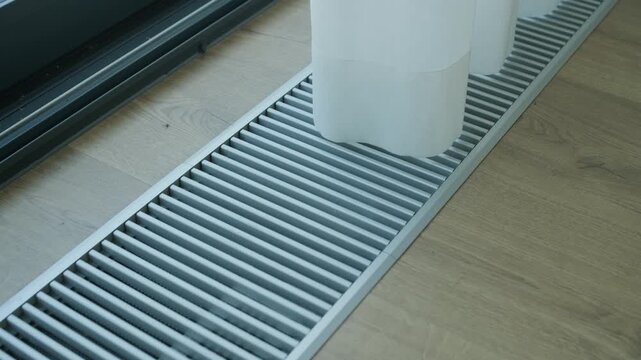 Metal floor vent grille near a large window on wooden flooring, part of a modern interior ventilation or heating system.