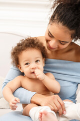 Smiling mother holding her cheerful baby boy on lap, both laughing and looking at camera. Warm family portrait full of love, tenderness, and happiness in bright cozy home.