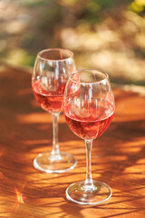 Two glasses of rose wine on wooden table with warm sunlight and dappled shadows in outdoor setting