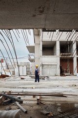 Fototapeta premium Man wearing hard hat standing on construction site surrounded by unfinished concrete building structures and exposed steel rebar, observing progress