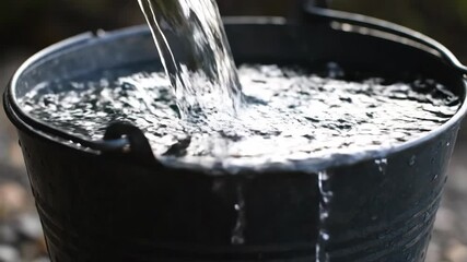 Clear Water Pouring into Black Bucket Overflowing with Splashes.