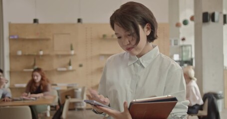 A young Asian businesswoman walks through a busy, diverse office while talking on her phone and holding a tablet. - Powered by Adobe