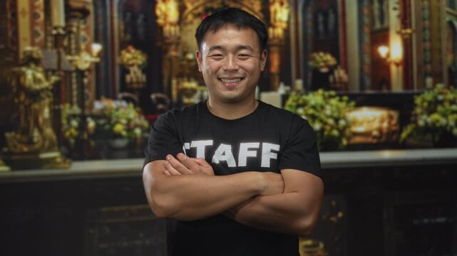 Young chinese man smiling and wearing staff tshirt crosses arms in church building; service faith devotion.