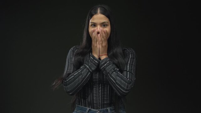 Young latin woman with hands covering mouth gesture in studio with dark green background; surprise.