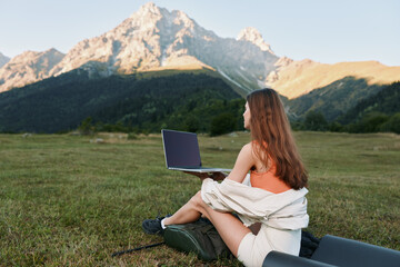 A relaxed woman sits on a blanket with a laptop in a scenic meadow. She wears casual attire and...