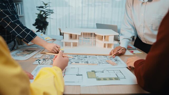 A diverse team of architects engages in a collaborative discussion at a modern office table, surrounded by blueprints, models, and construction plans, showcasing teamwork and creativity. SACTR - Powered by Adobe