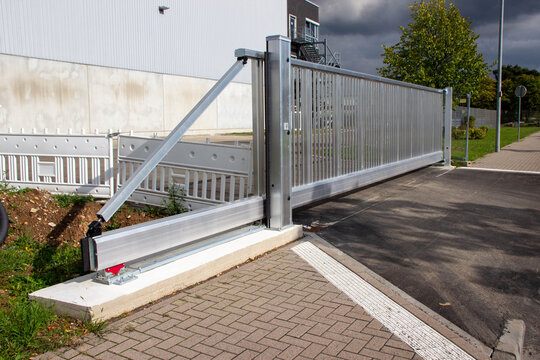 Modern sliding gate made of metal, partially open, showcasing its sleek design and functionality, set against an industrial backdrop with green landscaping